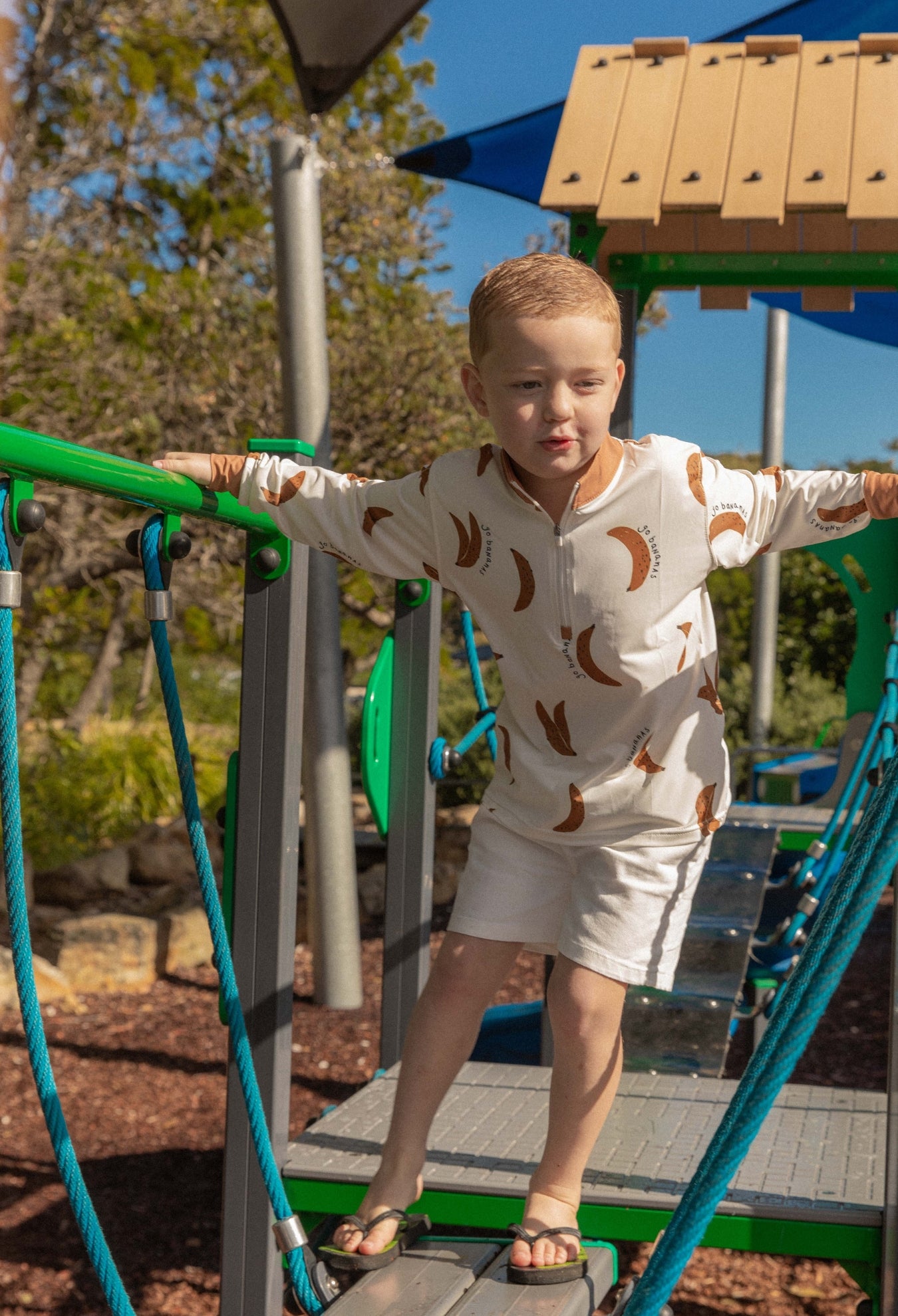 Child playing in UPF 50+ sun shirt on a playground structure with a clear sky and trees in the background